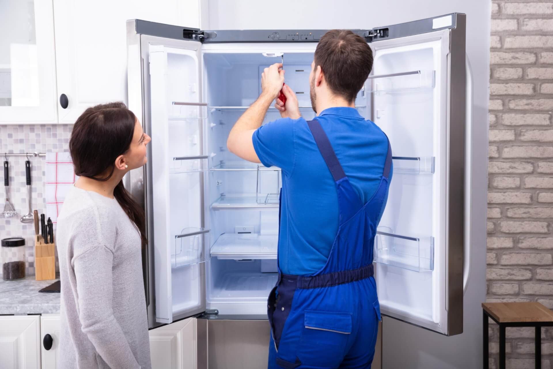 Professional appliance repair technician servicing a refrigerator while assisting a homeowner.